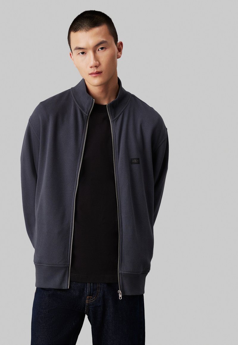 Dunkelgraue Zip-Jacke mit hohem Kragen und strukturiertem Material, kombiniert mit einem schwarzen Shirt, getragen über dunkelblauen Jeans.