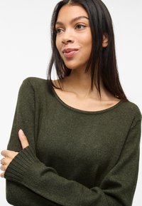 Maglione lavorato a maglia in verde scuro con scollo ampio, polsini a coste e texture morbida. Presenta una vestibilità morbida per un comfort ottimale.