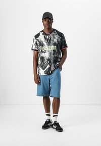 Majica s crno-bijelim tie-dye uzorkom i neon zelenim "adTUFF" logotipom, u kombinaciji s traper kratkim hlačama i crnim tenisicama s bijelim prugama na čarapama.