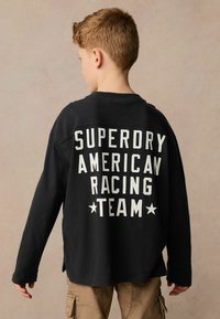 Tricou negru cu mânecă lungă, prezentând text alb "SUPERDRY AMERICAN RACING TEAM" pe spate, cu o croială lejeră și fante pe lateral.