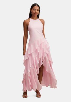 Donna che indossa un abito senza maniche color rosa pallido con orlo asimmetrico a strati e volant, e sandali con tacco alto e cinturini, in piedi davanti a uno sfondo neutro.