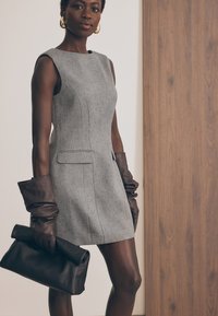 Sivá bezrukávová šaty s textúrovaným povrchom, so bočnými vreckami. V kombinácii s dlhými hnedými koženými rukavicami a čiernou klučkou.