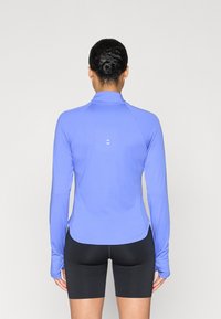 Top deportivo de manga larga color púrpura con cuello alto, fabricado con tejido elástico, que presenta un logo sutil en la parte trasera, combinado con pantalones cortos negros.