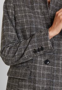 Blazer in grijs en zwart houndstooth patroon, met blauwe ruitaccenten, zwarte knopen en een gestructureerde stoffen afwerking.