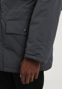 Szara kurtka puchowa o fakturowanej powierzchni, z kieszenią na klapę i guzikiem na rękawie. Zbliżenie na ramię i dłoń.