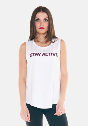Femme aux longs cheveux bruns portant un haut blanc sans manches avec le texte « STAY ACTIVE », associée à un pantalon noir, debout sur un fond blanc.