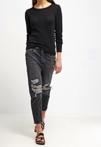 Kvinna i svart texturerad tröja, slitna svarta jeans och svarta sandaler med guldspännen, som går mot en vit bakgrund.