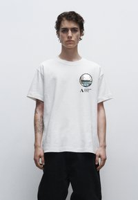 Hvid bomulds T-shirt med en rund grafisk print på venstre bryst, der viser et landskabsdesign og tekst. Løs pasform, korte ærmer, rund hals.