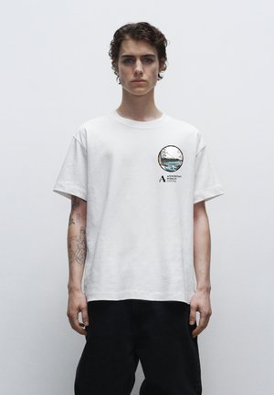 T-shirt en coton blanc avec un imprimé graphique rond sur la poitrine gauche, présentant un design de paysage et du texte. Coupe ample, manches courtes, col rond.