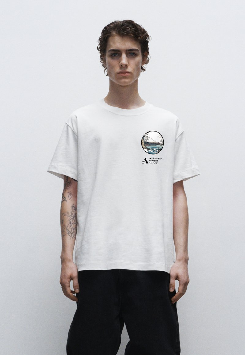 Weißes Baumwoll-T-Shirt mit rundem Grafikdruck auf der linken Brust, der ein Landschaftsdesign und Text zeigt. Lockere Passform, kurze Ärmel, Rundhalsausschnitt.