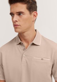 Beige polo shirt met een gestructureerde kraag, drie knopen en een klein patchzakje. Zachte stof met een egale afwerking.