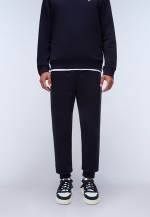 Uomo che indossa pantaloni della tuta neri, felpa nera e scarpe da ginnastica nere e bianche, in piedi davanti a uno sfondo chiaro e uniforme.