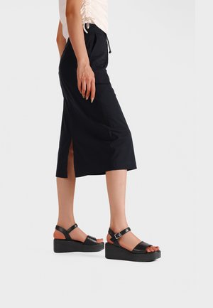 Donna con gonna midi nera con spacco laterale e sandali platform neri con cinturini alla caviglia, in piedi su uno sfondo chiaro.