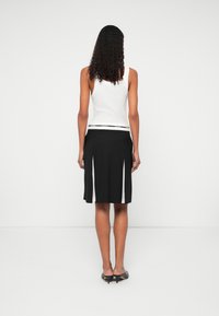 Kobieta z kręconymi włosami, ubrana w białą bluzkę bez rękawów, czarną spódnicę z białymi wstawkami i czarne szpilki, stojąca na tle jasnej, białej ściany.