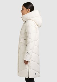 Geacă matlasată albă, cu guler înalt și glugă mare. Are un design matlasat, buzunare cu fermoar și un logo pe mânecă.