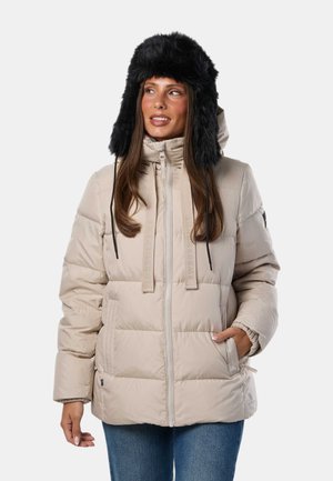 Femme portant une doudoune beige avec capuche et un chapeau trappeur noir en fourrure, regardant légèrement vers sa gauche avec la main gauche dans sa poche.