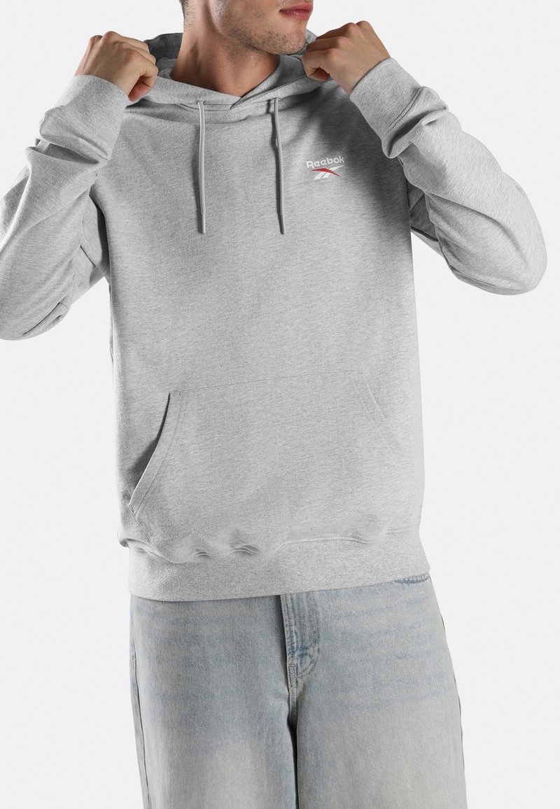 Mann, der die Kapuze eines schlichten grauen Reebok-Hoodies mit Fronttasche anpasst, trägt hellblaue Jeans, vor weißem Hintergrund.