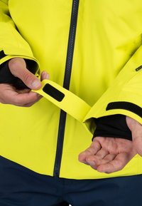 Chaqueta impermeable de color amarillo brillante con acentos en negro. Cuenta con puños ajustables con Velcro, tejido suave y diseño de cremallera frontal.