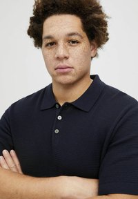 Tamsiai mėlynas polo marškinėlis, pagamintas iš tekstūruotos medžiagos, su klasikiniu apykakle, trijų mygtukų užsegimu ir trumpomis rankovėmis. Rankos sukryžiuotos priekyje.