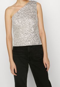 Silver sequin one-shoulder top with a fitted design. Smooth texture, metallic sheen, paired with black high-waisted jeans.