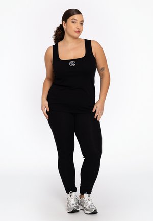 Débardeur noir à encolure carrée avec un petit logo sur la poitrine, associé à des leggings noirs et des baskets de sport argentées. Texture lisse.
