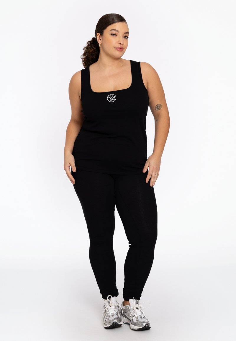 Zwarte tanktop met vierkante halslijn en klein logo op de borst, gecombineerd met zwarte leggings en zilveren sportschoenen. Gladde textuur.