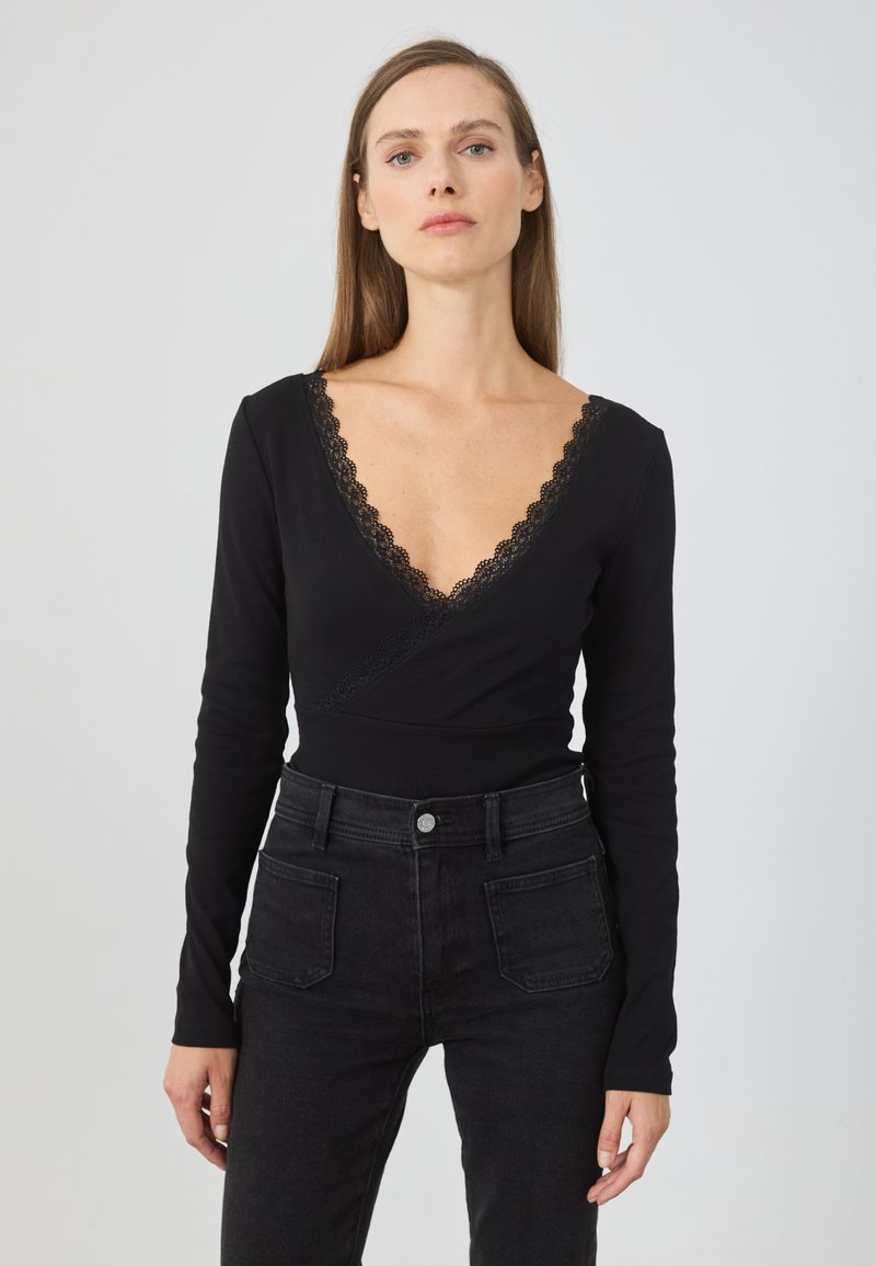 Sort langærmet top med dyb V-udskæring og blonder. Stof er ribbet og har en indpakningsdesign, der komplementerer tætsiddende sorte jeans.