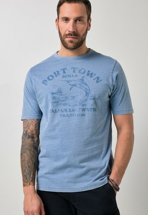 Brodaty mężczyzna z wytatuowaną lewą ręką w wyblakłej niebieskiej koszulce z napisem "Port Town Scilla Italian Saltwater Tradition".