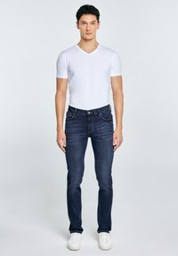 Mladić stoji, nosi bijelu majicu s V-izrezom, tamnoplave traperice i bijele tenisice, okrenut prema naprijed na jednostavnoj pozadini.