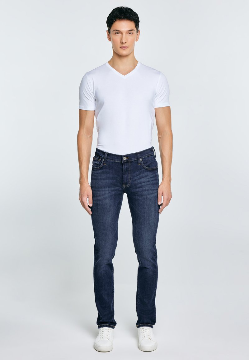 Mladić stoji, nosi bijelu majicu s V-izrezom, tamnoplave traperice i bijele tenisice, okrenut prema naprijed na jednostavnoj pozadini.