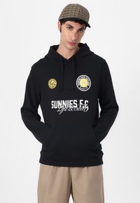 Čierna mikina s klokaniem vreckom, so žltými a bielymi logami na hrudníku a výrazným textom "SUNNIES F.C.". Materiál z bavlneného zmesi.