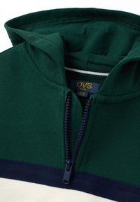 Sudadera para niños en verde y blanco con cremallera y detalles en azul marino, parcialmente cerrada, mostrando la etiqueta de OVS para niños en el interior del cuello.