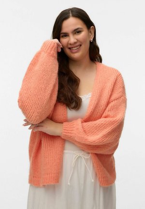 Junge Frau mit langen dunklen Haaren lächelnd, trägt eine locker gestrickte Pfirsichfarbene Strickjacke über einem weißen Oberteil und weißen Hosen mit Kordelzug, vor schlichtem Hintergrund.