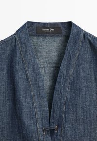 Veste bleu denim avec un col en V profond, des coutures marron visibles et une étiquette noire indiquant "Massimo Dutti Made in China" au niveau du col.