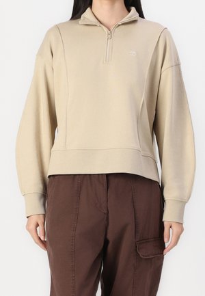 Osoba ubrana w beżową bluzę z krótkim suwakiem, z prążkowanymi mankietami, oraz brązowe spodnie bojówki z patką na kieszeni, stojąca na jednolitym tle.