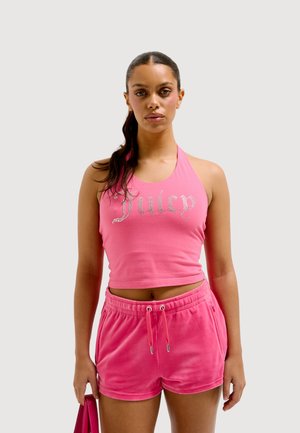 Jonge vrouw met donker haar in een paardenstaart, draagt een roze "Juicy" mouwloos crop top en bijpassende roze short, en houdt een roze tas vast.