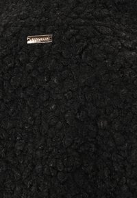 Schwarzer, strukturierter Stoff mit einer flauschigen, lockigen Oberfläche. Verfügt über ein kleines, metallisches Etikett, das mit dem Markennamen graviert ist.