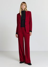 Completo rosso con blazer sartoriale e pantaloni a gamba larga, top nero strutturato sotto, bottoni evidenti e finitura liscia.