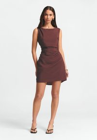 Abito aderente senza maniche in tessuto bordeaux scuro, con vita arricciata e orlo che cade sopra il ginocchio. Indossato con sandali neri.