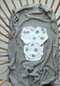 Weißer Langarm-Body für Babys mit blauen Wolkenmustern auf einer grauen Strickdecke, die auf einem geflochtenen Holzstuhl drapiert ist.