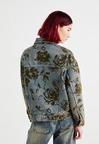 Jeans jakna s cvetnim temnozelenim vzorcem na svetlo modri podlagi. Klasičen kroj z gumbom spredaj in dvema stranskima žepoma.
