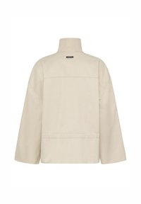 Beežika kõrge kaelusega jakk pikkade varrukatega, lihtsad õmblusdetailid ja väike must "IW.EDITION" silt ülaosas seljal.