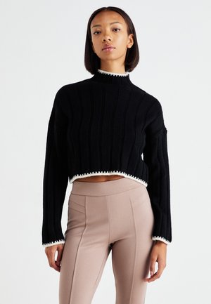 Black cropped sweater with ribbed texture, high collar, and white scalloped hem. Paired with beige fitted leggings.