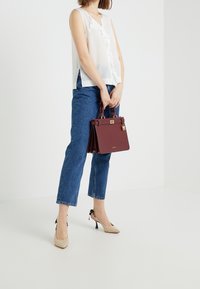 Biała bezrękawnikowa bluzka z falbanami, niebieskie proste dżinsy, beżowe obcasy oraz burgundowa torebka o strukturalnym kształcie ze złotymi elementami.