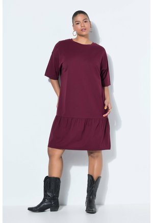 Mujer vestida con un vestido burdeos suelto hasta la rodilla con mangas cortas y botas vaqueras negras, de pie frente a un fondo claro.