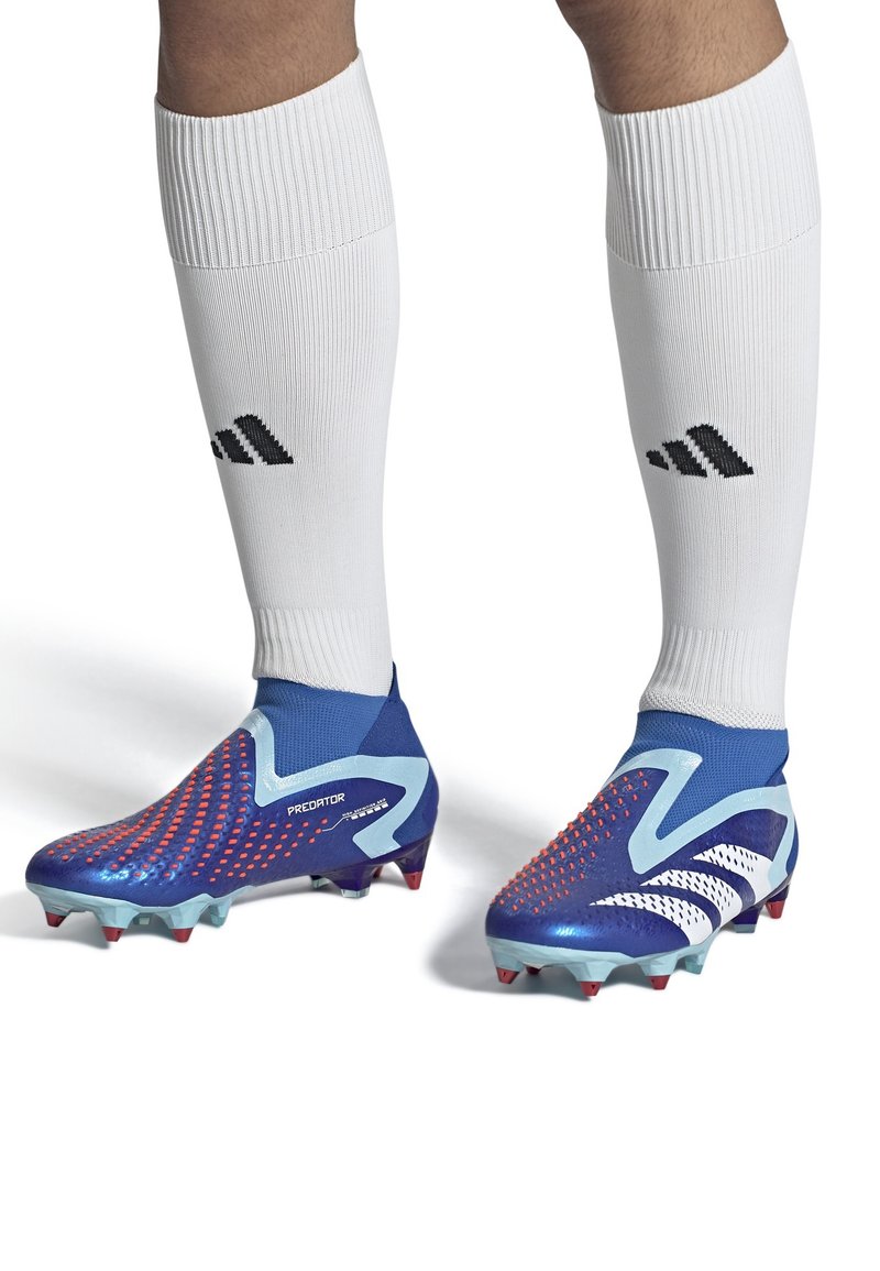 Blå fotbollsskor med röda prickmönster, vita accenter och en texturerad yta. Matchas med vita strumpor med en svart logotyp.