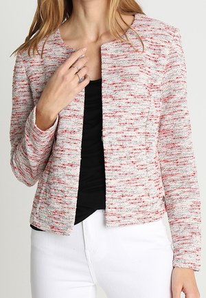 Femme portant une veste texturée blanche à manches longues avec des taches rouges sur un haut noir et un pantalon blanc, avec des bagues à la main gauche.