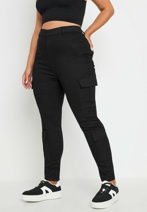 Femme portant un pantalon cargo noir taille haute avec poches à rabat et baskets noires et blanches, debout devant un fond clair uni.