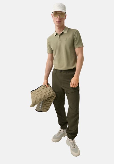 Olive polo shirt, dark green cargo pants, beige puffer bag, light grey sneakers, white cap, and oversized sunglasses on a neutral background.