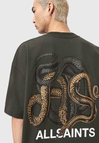 Oliwkowo-zielony oversize t-shirt z dużą czarno-złotą grafiką węża na plecach, z misternymi wzorami i napisem "ALLSAINTS".